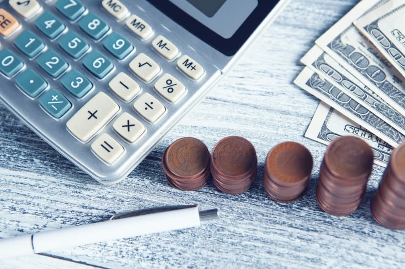 Calculator, stacks of coins showing growth, US hundred-dollar bills, and a pen on a wooden desk, representing revenue recognition and financial accounting